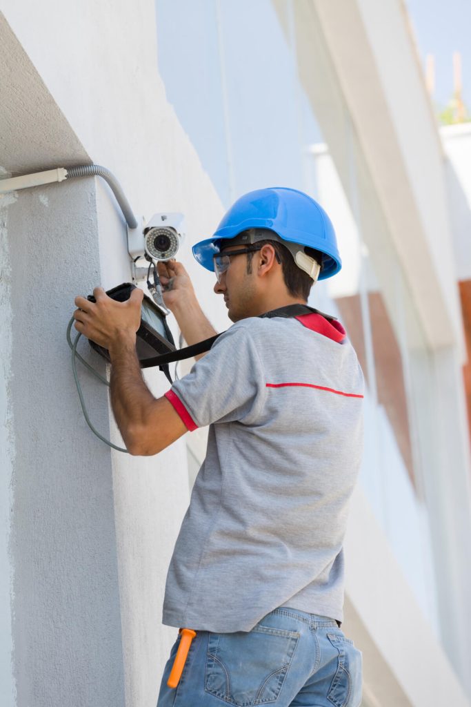 A worker wearing a blue safety helmet and gray shirt installs or repairs a security camera on an exterior wall, using tools and a device while standing on the ground.