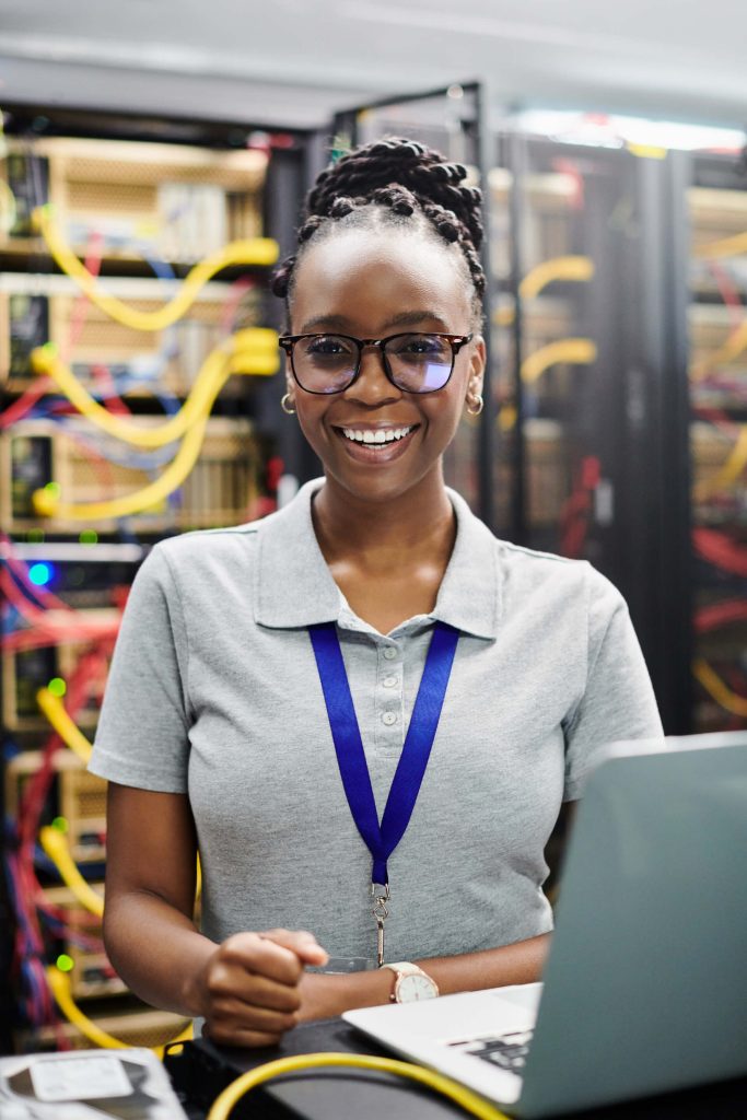 A smiling woman wearing glasses and a gray polo shirt stands in a server room with network cables and servers in the background. She has a lanyard around her neck and is working on a laptop.
