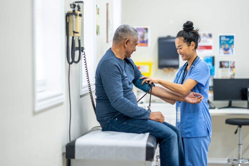 A nurse smiles while measuring the blood pressure of a seated man in a medical examination room. The man sits on an exam table and both appear to be enjoying a friendly interaction.