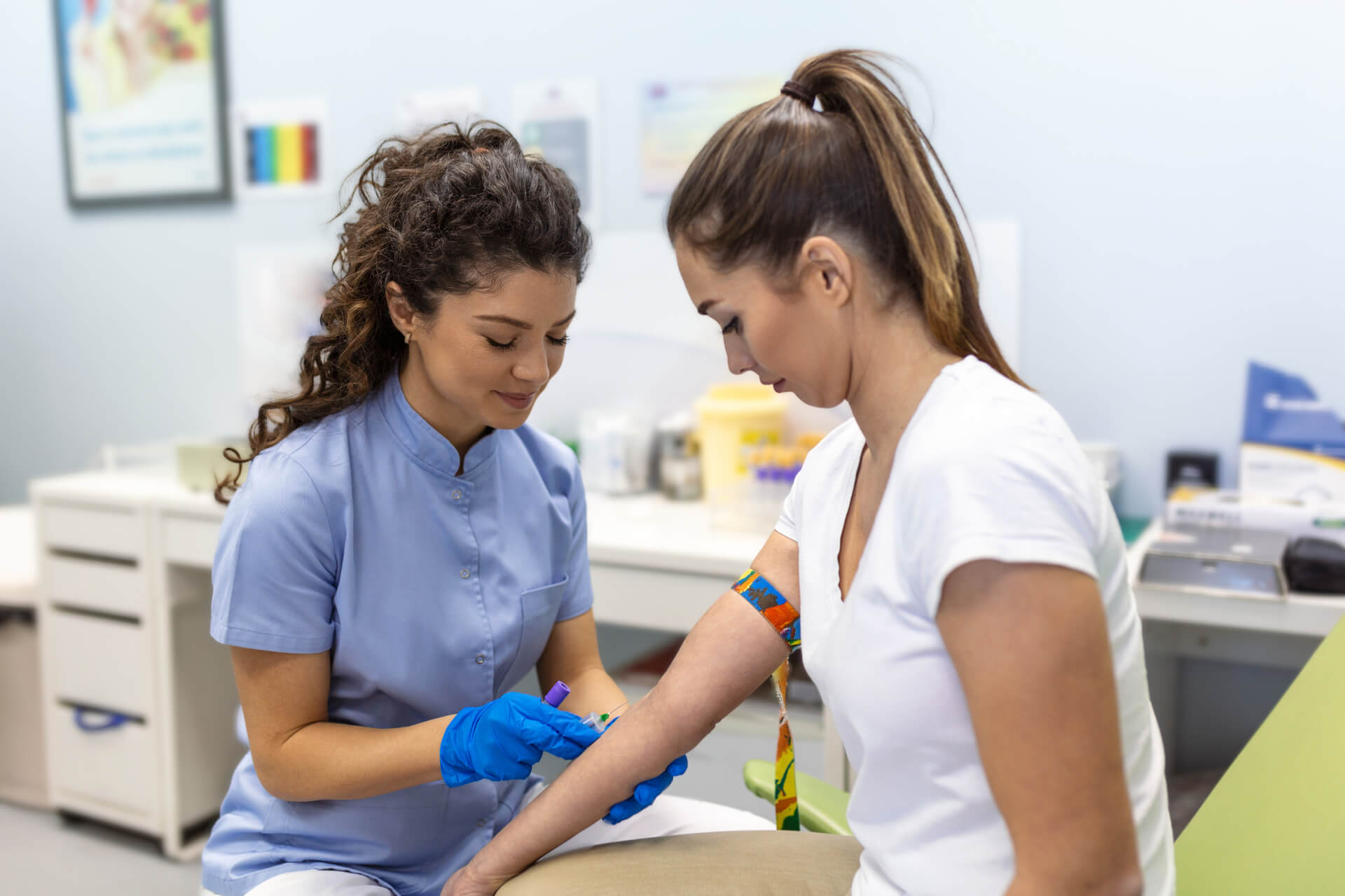 A healthcare professional wearing blue gloves prepares to draw blood from a seated woman’s arm in a medical clinic setting. The woman has a tourniquet on her arm and looks away as the nurse works.